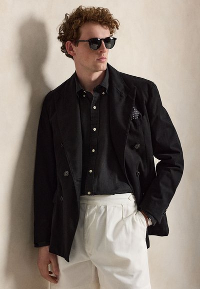 Άνδρας με σγουρά μαλλιά που φορά μαύρα γυαλιά ηλίου, μαύρο πουκάμισο με κουμπιά, μαύρο σακάκι με μαντηλάκι με πουά και λευκό πλισέ παντελόνι.