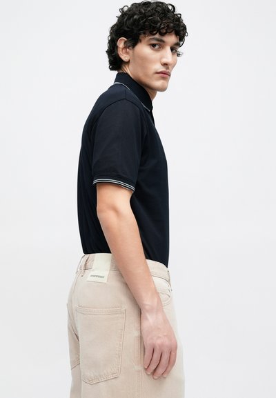 Μπλε ναυτικό πόλο με λευκές ρίγες στον γιακά και τις μανσέτες, σε συνδυασμό με ανοιχτό μπεζ παντελόνι που διαθέτει πίσω τσέπες και ετικέτα μάρκας.