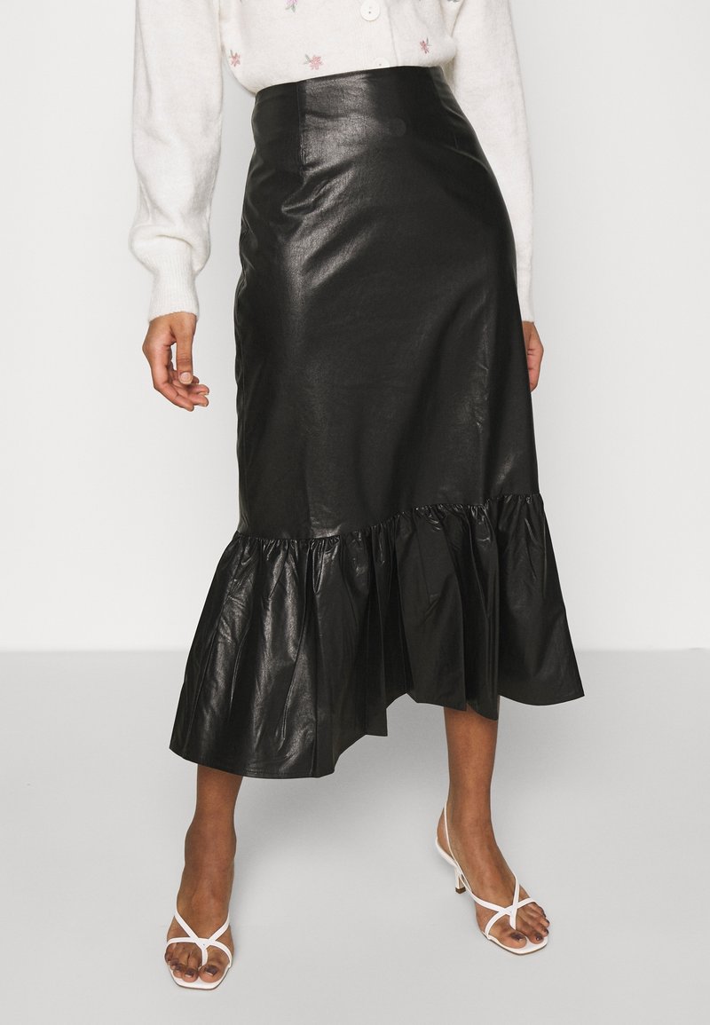 Femme portant une jupe midi asymétrique en similicuir noir avec un ourlet à volants, un pull blanc brodé de fleurs et des talons blancs à lanières.