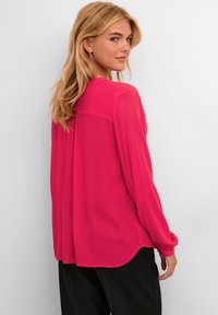 Blusa de manga larga en rosa brillante, confeccionada con tejido ligero, que presenta un respaldo pliegue y un dobladillo redondeado para un ajuste relajado.