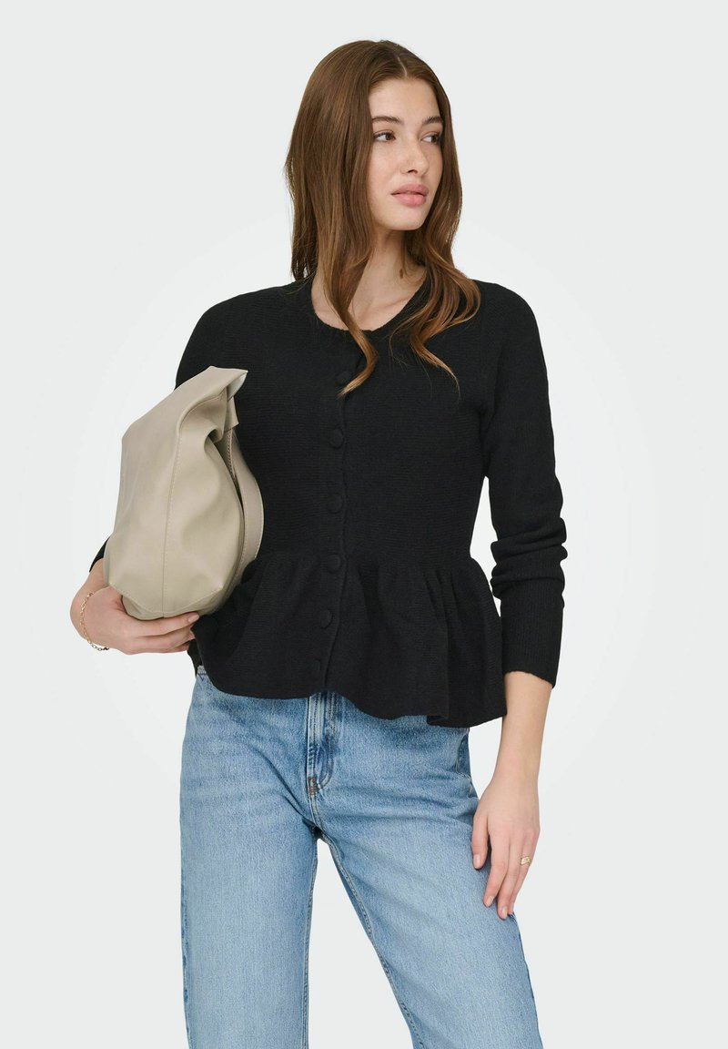 Sort strikket cardigan med peplumkant, med knapper foran. Kombineret med lyseblå højtaljede jeans og en beige taske.