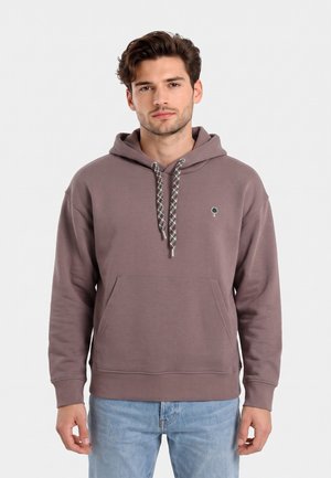 Joven con cabello castaño que lleva una sudadera con capucha color malva con cordones estampados y vaqueros azul claro, de pie frente a un fondo blanco liso.