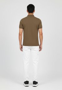 Camisa polo marrón de mangas cortas, combinada con pantalones cargo blancos y zapatillas negras, vista desde atrás contra un fondo liso.