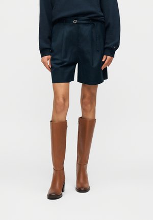 Mujer con pantalones cortos azul marino con cinturón negro, top de manga larga negro y botas altas de cuero marrón con tacón, de pie frente a un fondo liso.