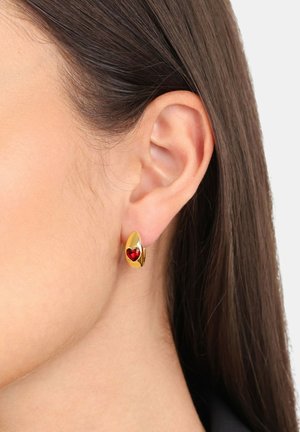 Pendientes de color dorado con forma de lágrima, que presentan una gema en forma de corazón rojo en el centro, exhibiendo un acabado suave y brillante.