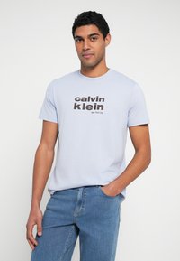 Helehall sinine puuvillane t-särk, millel on julge tumepruun "calvin klein" logo ja tekst "New York City". Lühikesed varrukad ja ümmargune kaelus.