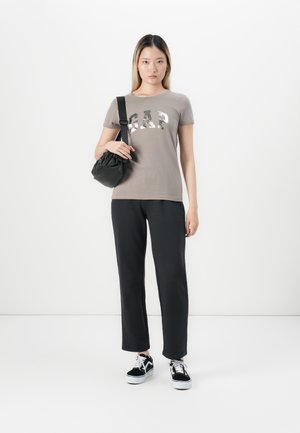 Jeune femme debout, portant un t-shirt taupe de GAP, un pantalon noir, des baskets noir et blanc, et portant un sac à bandoulière noir.