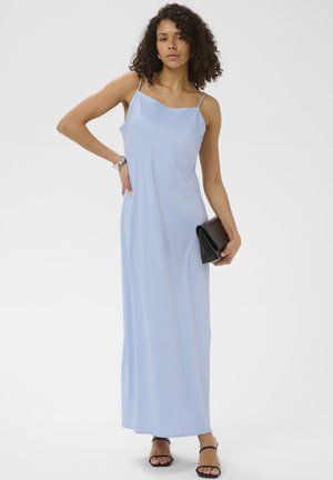 Femme aux cheveux bouclés portant une robe longue sans manches bleu clair, des sandales noires à lanières, un bracelet en argent, et tenant une pochette noire.