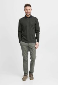 Mørkegrønn langermet polo skjorte med svart krage, med en liten logo på brystet. Kombinert med lyse grå bukser og brune sko.