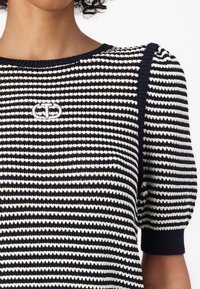 Pruhovaný pletený top v námořnické modré a bílé barvě s kulatým výstřihem, krátkými nabíranými rukávy a výšivkou loga na hrudi.