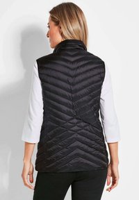 Schwarze, gesteppte Weste mit einem Chevron-Muster. Hoher Kragen, ärmelloses Design und glatte Textur, getragen über einem weißen Langarmshirt.