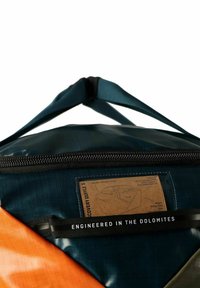 Bolsa de lona en azul verdoso y naranja con acabado brillante, que presenta una cremallera grande, etiqueta que detalla el material y correas ajustables.