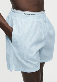 Άτομο με σκουρόχρωμη επιδερμίδα φοράει ανοιχτό μπλε υφασμάτινες βερμούδες με ελαστική μέση, χέρι στην τσέπη και ορατή μέση εσωρούχου Calvin Klein από πάνω.