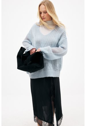 Kvinde iført en lyseblå strikket sweater og sort nederdel med blonder, der holder og kigger ned i en stor sort håndtaske.