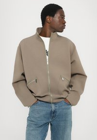 Chaqueta beige con cremallera y cuello alto, dos bolsillos frontales y corte relajado, combinada con jeans azules. Textura de tela suave.