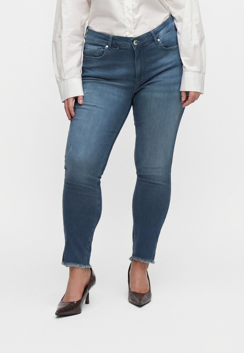 Donna che indossa jeans slim fit blu medio con orli sfrangiati, abbinati a una camicia bianca con bottoni e décolleté scuri a punta.