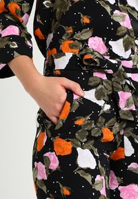 Blommönstrat tyg med rosa, vita och orange rosor på svart bakgrund; har ett bälte och en framficka. Texturerat material synligt.
