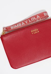 Rode leren pouch met textuurafwerking, gouden logo "BIMBA Y LOLA," en een afneembare schouderband met een herhalend patroon in het wit. Ritssluiting.