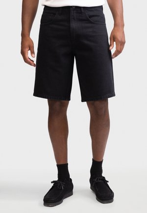 Man draagt zwarte denim shorts, zwarte sokken en zwarte veterlaarzen en staat tegen een effen achtergrond.