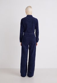 Marine corduroy jumpsuit met een aansluitend lijfje, lange mouwen en een wijd uitlopende broekspijp. Heeft achterzakken en een ceintuur in de taille voor extra vorm.