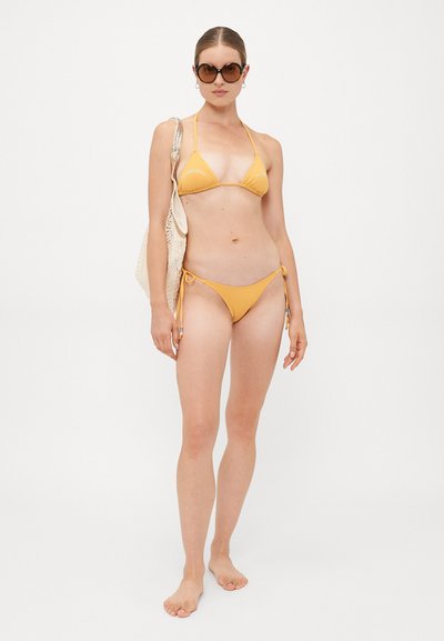 Frau in gelbem Bikini, mit übergroßen braunen Sonnenbrillen, die eine beigefarbene Netztragetasche trägt, barfuß vor weißem Hintergrund stehend.