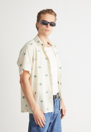 Short-sleeve beige shirt with green floral embroidery, paired with a white tank top and light blue jeans. Black rectangular sunglasses worn.