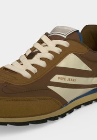 Καφέ και μπεζ sneaker Pepe Jeans με λευκά κορδόνια, πάνελ από σουέντ και δέρμα και λαστιχένια σόλα με ορατό πέλμα.