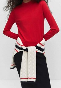 Haut rouge à manches longues associé à un pull tricoté blanc et bleu marine à rayures noué autour de la taille, contrastant avec une jupe noire.