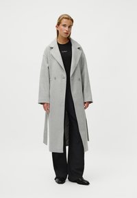 Cappotto di lana grigio chiaro con ampi rever, bottoni doppi e maniche lunghe, indossato sopra un top nero e pantaloni scuri larghi.