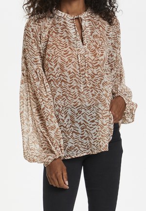 Femme portant un chemisier beige et marron à manches longues avec des poignets froncés et un décolleté en forme de goutte, associé à un pantalon noir.