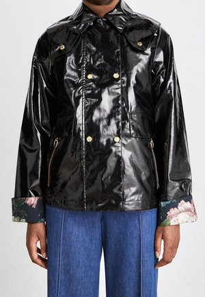 Veste noire brillante à double boutonnage avec boutons dorés, poignets intérieurs fleuris et poches latérales zippées portée sur un jean bleu et un col roulé marron.