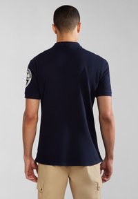 Námorně modré polo tričko vyrobené z bavlny s krátkými rukávy. Na levém rukávu se nachází kulatý logový nášivka. Klasický design límce.