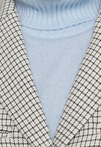 Beige och svart houndstooth-mönstrad blazer över en ljusblå ribbad polotröja. Texturerade tyger med distinkta detaljer på krage och slag.