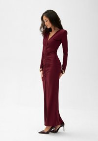 Dlouhá vínová šaty s dlouhými rukávy, výstřihem do V a nabíráním podél předního dílu. V páru s lesklými vysokými podpatky s špičatou špičkou.
