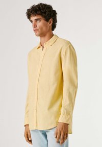 Homme aux cheveux bouclés et foncés portant une chemise boutonnée à manches longues jaune pâle et un jean bleu clair, debout devant un fond uni.