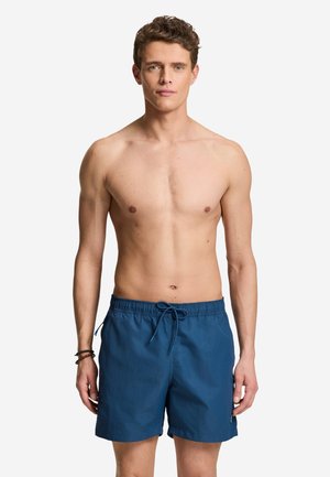 Jeune homme debout, torse nu, portant un short de bain bleu avec un cordon, et des bracelets marron à son poignet gauche, face à l'avant.