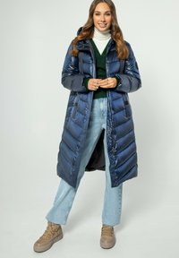 Giacca puf blu navy con design trapuntato, chiusura a zip e cappuccio. Abbinata a un maglione verde e denim blu chiaro, completata da sneakers beige.