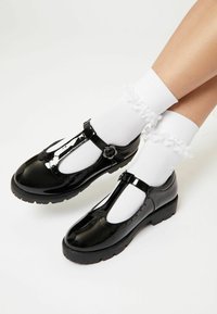 Zapatos de charol negro tipo T con punta redondeada, adornados con una hebilla, acompañados de calcetines blancos con volantes. Suela de goma para mayor agarre.