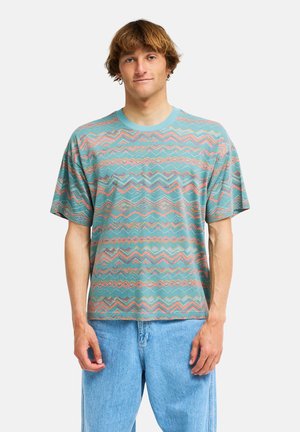 Jonge man met kort bruin haar, gekleed in een turquoise T-shirt met patroon en lichtblauwe spijkerbroek tegen een effen witte achtergrond.