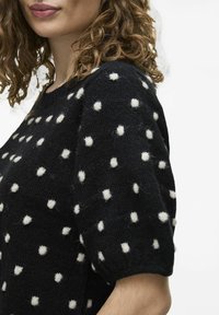 Melna adīta džemperis ar baltām teksturētām punktiņām, īsām piedurknēm un apaļu dekoltē, izskatās mīksts un ērts.