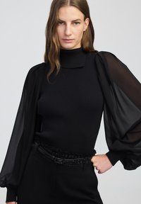 Maglia nera a coste con maniche a sbuffo trasparenti, caratterizzata da un collo alto e un colletto sottile. Abbinata a una cintura nera con texture.