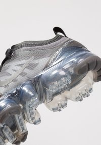 Sneaker grigio a rete con tomaia testurizzata, dotato di una suola ad aria trasparente, lacci prominenti e un accento del logo swoosh riflettente.