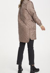 Prošívaná béžová bunda s vysokým límcem, bočními zipovými kapsami a žebrovanými manžetami, spárovaná s černými džíny a kotníkovými botami.