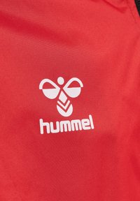Röd tyg med en jämn textur har en vit logotyp bestående av rundade former och texten "hummel" under den.