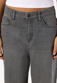 Vyötärö ja lantio henkilöllä, joka pukeutunut harmaisiin korkeavyötäröisiin farkkuihin, joissa edessä nappikiinnitys, ja valkoiseen lyhyt pusero, kädet rentoina sivuilla.