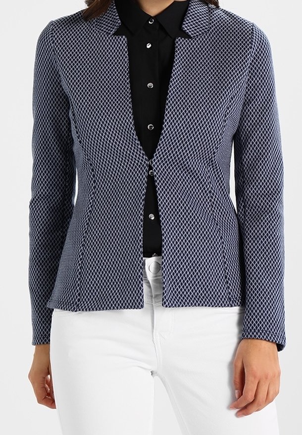 Femme portant un blazer ajusté bleu marine et blanc à motifs, sur une chemise noire boutonnée, associée à un pantalon blanc.