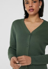Cárdigan de punto verde con cuello en V y dos cierres de botón. Mangas largas con puños acanalados. Textura suave y tejido de peso medio.