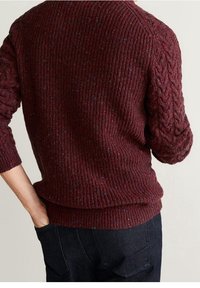 Pull en tricot bordeaux avec une texture côtelée, des manches à torsades et des taches multicolores partout. Porté avec un pantalon foncé.