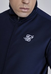 Mornarica zip-up jakna s visokim ovratnikom, izrađena od glatkog materijala. Ima bijeli izvezeni logo na prsima i metalni patentni zatvarač.