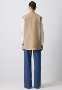 Beige mouwloos vest met een kraag, gecombineerd met losse blauwe denim broek. Strakke witte shirtmouwen zichtbaar, wat een gelaagde uitstraling creëert.